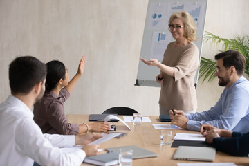 Smiling indian businesswoman raising hand asking mature mentor in boardroom at company meeting. Diverse happy woman employee in conversation asks older lead question about business sit at table.