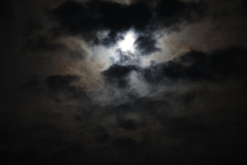 Mystical landscape of the night sky.The outline of the full moon is hidden by black and gray fluffy clouds on a smoky background of voluminous haze that hides the light.A mix of dark tones