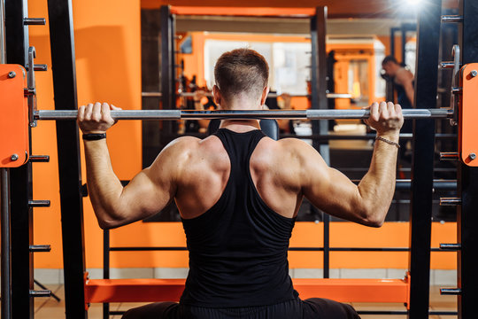 Average Plan, Back View. A Muscular Man Dressed In Black Sportswear Is Training Muscles, Biceps And Triceps - With An Effort He Lifting The Bar. Fitness And Sport. Healthy Lifestyle Concept