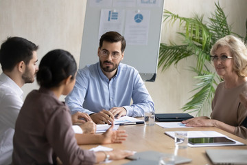 Serious businessman listening indian employee about project sit at table in boardroom at company meeting. Confident leader discuss business strategy with diverse woman and colleagues.