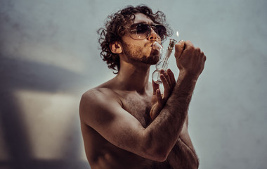 Young caucasian male model is naked and isolated on the grey background, holding his tiny glass bong