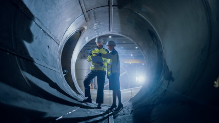Two Heavy Industry Engineers Walking Inside Pipe, Use Laptop, Have Discussion, Checking Design. Construction of the Oil, Natural Gas and Biofuels Transport Pipeline. Industrial Manufacturing Factory © Gorodenkoff