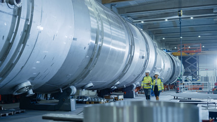 Two Heavy Industry Engineers Walking Through Pipe Manufacturing Facility, Use Digital Tablet, Have...