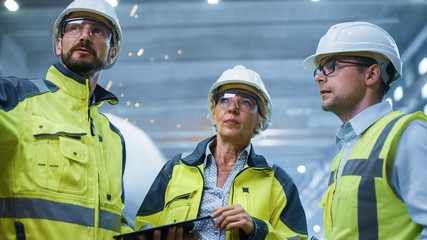 Three Heavy Industry Engineers Stand in Pipe Manufacturing Factory, Use Digital Tablet Computer,...