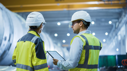 Two Heavy Industry Engineers Stand in Pipe Manufacturing Factory, Use Digital Tablet Computer, Have...