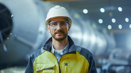 Portrait of Smiling Professional Heavy Industry Engineer / Worker Wearing Safety Uniform, Goggles and Hard Hat. In the Background Unfocused Large Industrial Factory where Welding Sparks Flying - obrazy, fototapety, plakaty