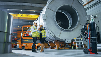 Two Heavy Industry Engineers Stand in Pipe Manufacturing Factory, Use Digital Tablet Computer, Have...