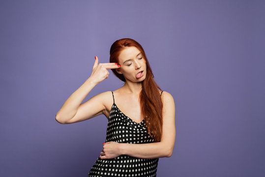 A red head girl with fingers at the temple . A woman is bored on the purple background. A girl with stiking out tongue