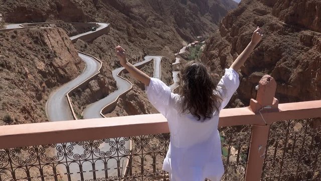 Back view of tourist woman in white dress raising hands up and enjoy view of windy mountain road in the Dades Gorge, Gorges Du Dades, Morocco