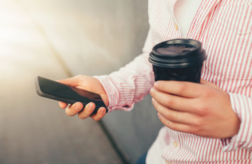 Close-up of smartphone and black disposable cup of coffee or tea in male hands. Selective focus