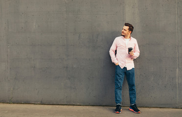 Obraz na płótnie Canvas A young stylish smiling man in blue jeans and striped pink shirt drinking coffee leaning against the concrete grey wall on the street. City lifestyle. Copy space