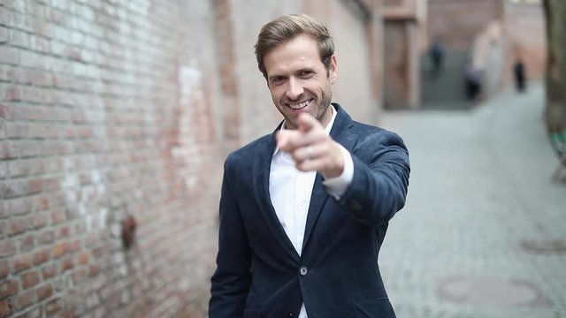 sexy businessman standing with one hand in pocket near a brick wall and pointing forward