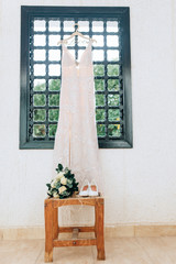white wedding dress in the window with the wooden table, wedding bouquet and white shoes.