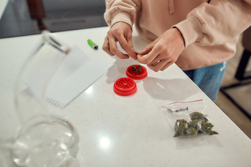 Experience. Cropped shot of woman preparing cannabis buds for grinding using red marijuana grinder, while standing in the kitchen. Glass water pipe or bong on the table