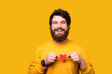 Handsome smiling man is holding a small red heart over his chest on yellow background.