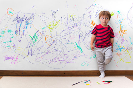 Child mischief. Boy with a distracted face because he drew the entire wall. Little boy leaning against the white wall where he made many drawings with colored pencils. Kid indoors, at home.