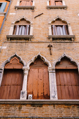 Fototapeta na wymiar Windows of Venice are a building of several floors, made of red brick, three windows with brown shutters at bottom, two windows with wooden shutters-screen, stone columns and a sharp top by window