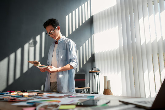 Working On A New Project. Smiling Asian Man In Eyeglasses Looking At Color Palette While Standing Near The Office Table With A Lot Of Creative Stuff On It