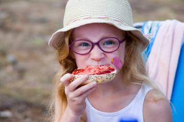 Rothaariges Mädchen mit Sonnenhut und Brille isst ein Mohnbrötchen mit Marmelade