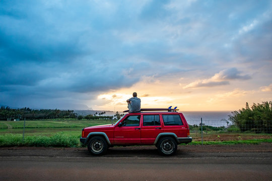 A young man sitting on top of his vehicle watching a colorful sunset in Hawaii.