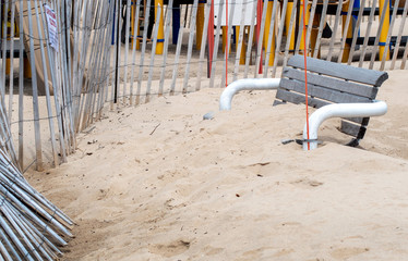 Park bench covered in sand next to a fenced off empty playground.  everything under lock down...