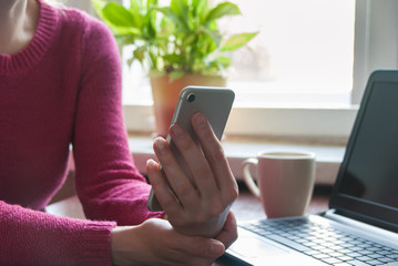 Close up of a woman using smartphne on kitchen table with coffee Mug and laptopNewspaper and Checking Cell Phone Messages on Relaxing Morning at Home