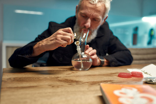 Seeking relaxation. Senior man lighting cannabis in the bowl of glass water pipe or bong in the kitchen. Marijuana tools on the table. Legalization and dependence on light drugs concept