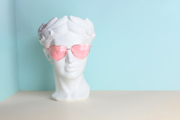 White sculpture of an antique head in pink glasses with hearts. On a geometric background of two colors.