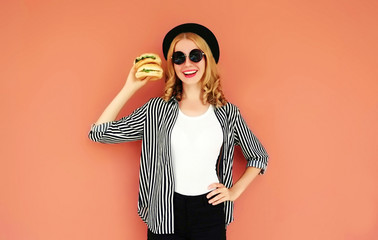 Portrait smiling young woman showing a burger wearing a black hat, sunglasses over wall background