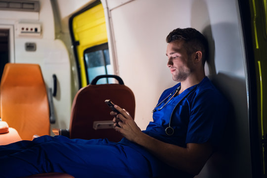 Young paramedic sitting with his smartphone in the ambulance car