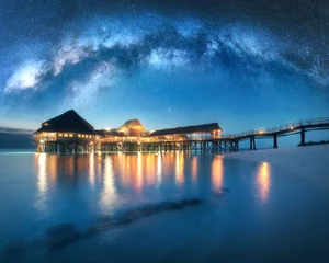 Poster Milky Way over wooden bungalow on the water in summer starry night. Landscape with hotel on the sea, illumination, jetty, sandy beach, sky with stars, reflection in water in Zanzibar, Africa. Space © den-belitsky