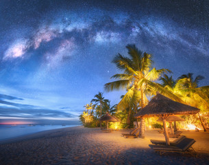 Voie lactée sur la plage de sable avec palmiers et transats et parasols la nuit en été. Paysage avec côte de la mer, beau ciel étoilé bleu, galaxie, palmiers verts. Voyage à Zanzibar, en Afrique. Espacer