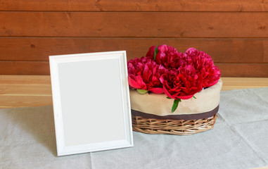 Red peony flowers in wicker basket and white frame on wooden table. Selective focus. View with copy space.