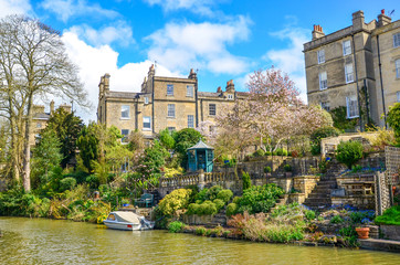 Historic houses and beautiful gardens with green plants and flowers by Canal river on a sunny spring day. Boat parked on the heritage waterway - Kennet and Avon Canal, Bath/United Kingdom.