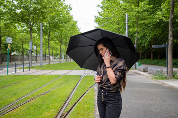 Une jolie femme au téléphone maintenant un parapluie noir sous la pluie à côté des rails de tramway dans la ville de Grenoble