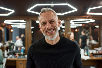 Fototapeta premium Happy handsome middle-aged man in barbershop. Portrait of elegant bearded man looking at camera and smiling while visiting barber shop or salon