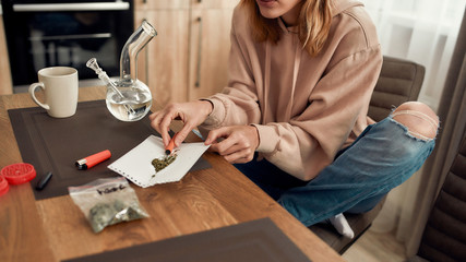 Way of improving your mood. Cropped shot of young caucasian woman putting weed in a tube while sitting in the kitchen. Bong, buds in a plastic bag, marijuana grinder and lighter on the table