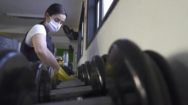 woman worker disinfects gym fitness equipment from coronavirus covid-19 with antibacterial sanitizer sprayer on quarantine. Cleaner in protective mask cleans training apparatus at workout area.
