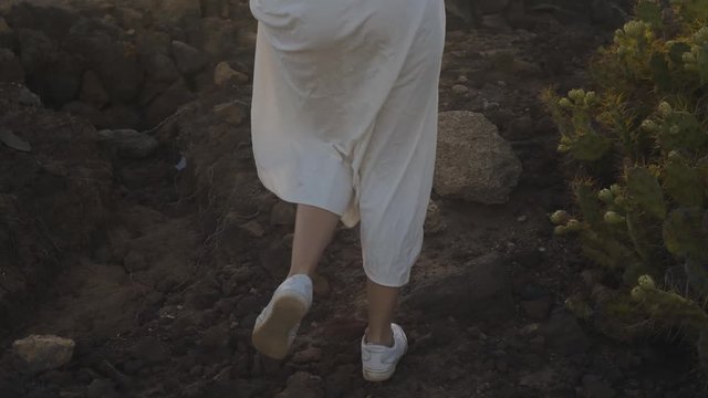 A girl walks along the summer island during sunset. Legs of a girl in beauties and a long white dress walk on mountains, stones. Atmospheric video