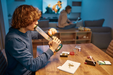 Device for smoking marijuana. Young guy lighting bong with cannabiswhile sitting in the kitchen at...