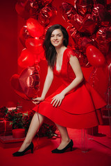 Woman with Hearts Balloons on a red background. Beauty Girl on Valentine's Day. Model in a stylish red dress.