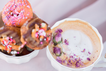 Romantic cup of coffee with editable dried rosebud flowers on top with mini donuts on pink table cloth