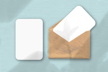An envelope with two sheets of textured white paper on a blue table background. Mockup with an overlay of plant shadows. Natural light casts shadows from a tropical plant.