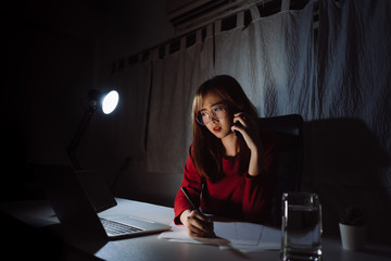 Asian woman working late time at night talking with smartphone, laptop computer home office unhappy emotion, Freelancer stay home business sleepless quarantine crisis coronavirus in dark room.