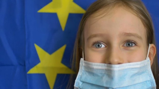 Close-up portrait of blond little girl with big blue eyes in disposible mask for protection of virus on European Union flag banner background in studio. Personal protective equipment