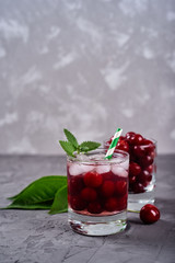 Fresh cherry lemonade with ice, mint and paper straw in sparkling glasses on gray table background, copy space. Cold summer drink. Berry cocktail