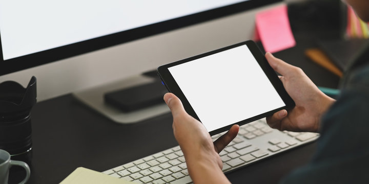 Cropped image of smart man hands holding a white blank screen computer tablet in front white blank screen computer monitor at the black working desk.