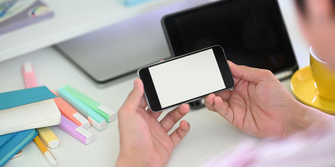 Cropped image of smart man's hands holding a white blank screen smartphone while sitting in front of computer tablet that putting on white working desk that surrounded by office equipment.