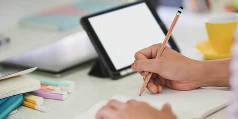Cropped image of young student hands writing on notebook while sitting in front of white blank screen computer tablet that putting on white student desk. Student doing homework at home concept.