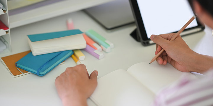 Cropped image of young student hands writing on notebook while sitting in front of white blank screen computer tablet that putting on white student desk. Student doing homework at home concept.